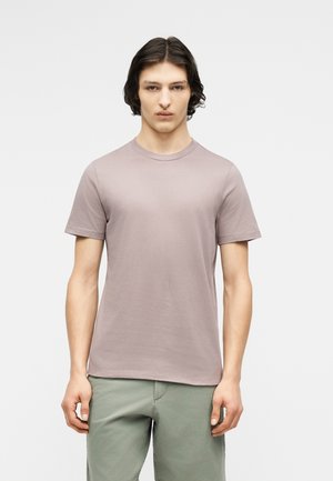 Νεαρός άνδρας με μεσαίου μήκους σκούρα μαλλιά, φορώντας μονόχρωμο ανοιχτό μωβ T-shirt και ανοιχτό πράσινο παντελόνι, στέκεται μπροστά σε απλό φόντο.