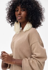 Beżowa bluzka z długim rękawem z delikatnym połyskiem, z szerokimi rękawami i okrągłym dekoltem. Duże białe kolczyki z frędzlami dodają szczegółów.