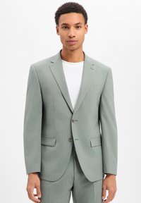 Giovane uomo che indossa una giacca e pantaloni abbinati grigio chiaro su una maglietta bianca a girocollo, in piedi davanti a uno sfondo bianco.