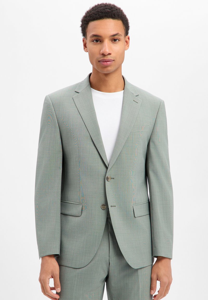 Giovane uomo che indossa una giacca e pantaloni abbinati grigio chiaro su una maglietta bianca a girocollo, in piedi davanti a uno sfondo bianco.