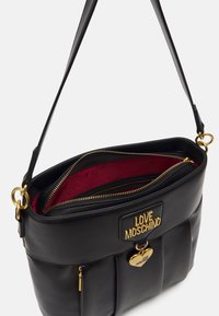 Bolso de mano de cuero sintético negro con un logo de tono dorado, charm en forma de corazón, interior rojo y compartimentos con cierre. Cuenta con una única correa para el hombro.