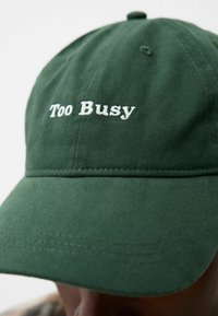 Casquette en coton verte au visière incurvée, arborant sur le devant le texte brodé en blanc "Trop occupé". Le tissu a une texture lisse.