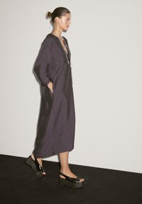 Langes, dunkelgraues Kleid mit weiten Ärmeln, einem tiefen Ausschnitt und seitlichen Taschen. Kombination mit glänzenden schwarzen Plateausandalen.