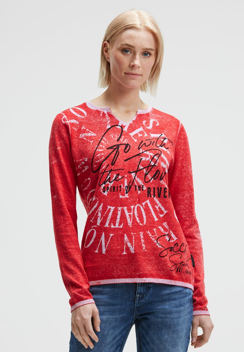 Langärmliges rotes Hemd aus weichem Stoff, mit weißen und schwarzen Textdrucken. Der Ausschnitt ist in zartem Rosa eingefasst. Kombiniert mit blauen Jeans.