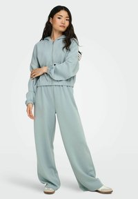 Frau in hellblauem, oversized Reißverschluss-Hoodie und passenden weit geschnittenen Hosen, die mit verschränkten Armen steht, weiße Sneaker sichtbar.