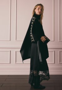 Fekete poncsó csillagmintás szegéllyel, hosszú fekete ruhával, amely csipkés szegéllyel rendelkezik, fekete bokacsizmával kiegészítve.