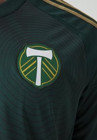Groene sporttrui met een cirkelvormig logo met een witte bijl en gele accenten, tegen een gestructureerde, gestreepte stof achtergrond.