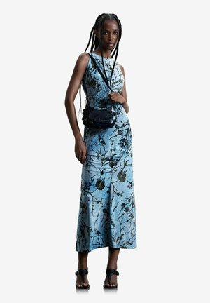 Naine pikkade patsidega, kes kannab sinist lillelist varrukateta midi kleiti, musta risti üle õla kandvat kotti ja musti kontsaga sandaale, seistes valge tausta ees.