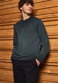 Dunkelgrünes langärmeliges Hemd mit Henley-Ausschnitt, drei Knöpfen und weicher Textur; schwarze Hose; Holz Hintergrund.