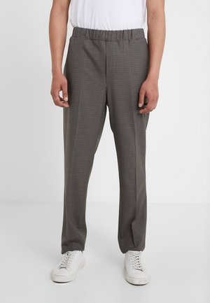 Man wearing brown and black checkered elastic waist pants paired with white sneakers and a white shirt, standing on a white floor.