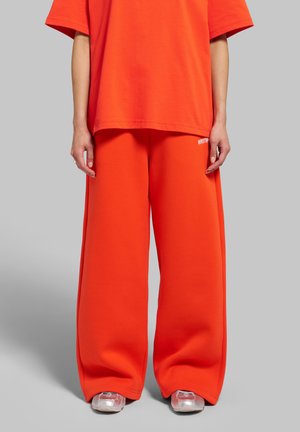 Personne portant un pantalon ample orange vif et une chemise orange assortie, debout sur un fond gris avec des chaussures transparentes visibles.