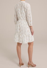 Kvinna iförd en vit blommig knälång klänning med 3/4 ärmar och beige klacksandaler, stående mot en neutral bakgrund.