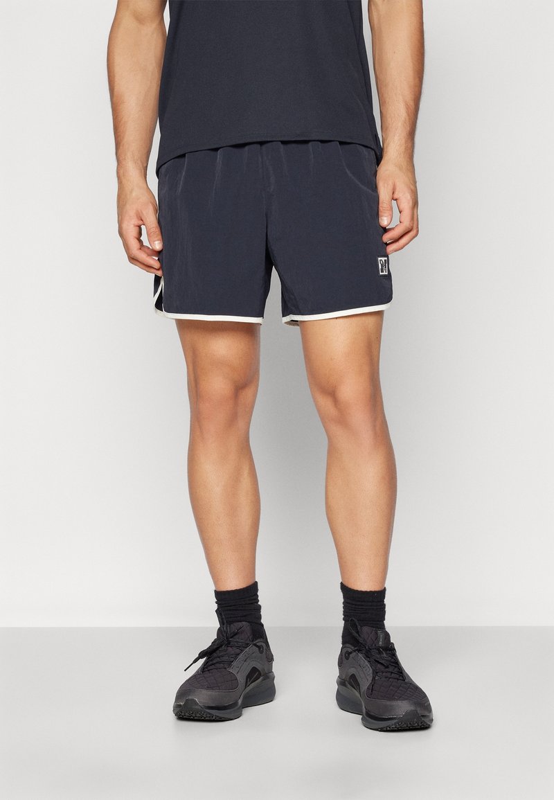Donkerblauwe zwemshorts met witte rand, elastische tailleband en zijzakken. Gedragen met zwarte sportschoenen en sokken.