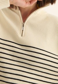 Pull-over crème en maille avec col à fermeture éclair sur un quart, présentant des rayures noires horizontales sur la poitrine et une texture côtelée sur toute la surface.