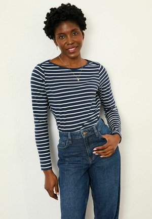 Kvinde med kort krøllet hår iført navy og hvidstribet langærmet skjorte og højtaljede blå jeans, smilende og let lænende.