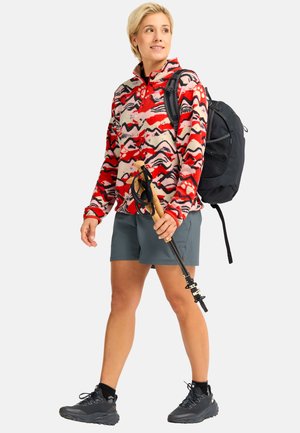 Femme en polaire rouge à motifs, short gris, chaussures de randonnée noires, portant un sac à dos noir et tenant des bâtons de trekking pliés, regardant vers la droite.