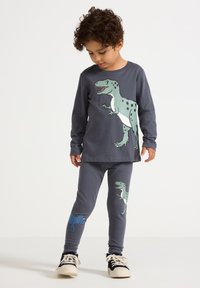 Bambino che indossa un maglione a maniche lunghe grigio scuro con stampa di dinosauri e leggings abbinati, con scarpe da ginnastica nere e bianche, che guarda in basso.