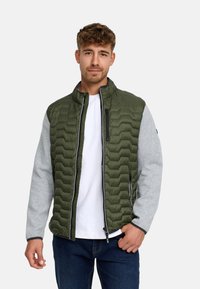 Ung mand iført en grøn quiltet vest med grå ærmer, hvid t-shirt og mørke jeans, stående mod en ensfarvet lys baggrund.