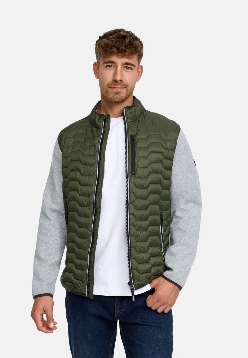 Ung mand iført en grøn quiltet vest med grå ærmer, hvid t-shirt og mørke jeans, stående mod en ensfarvet lys baggrund.