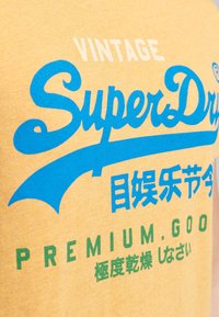 Camiseta de algodón amarilla con texto impreso en azul y verde, que incluye "Vintage SuperDry" y varios caracteres chinos, con un diseño clásico.