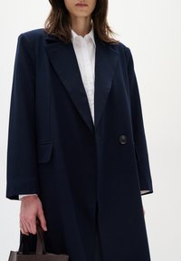 Cappotto lungo blu navy con silhouette strutturata, rever a punta, chiusura con un bottone e tasca. Indossato sopra una camicia bianca con colletto.