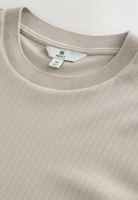 Lys beige ribbet trøje med rund hals, der har en glat tekstur og diskrete linjer. Etiketten er mærket "NEXT" med størrelse "M."