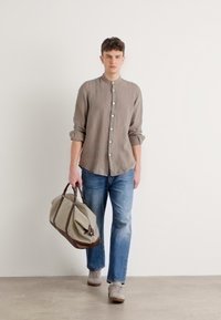 Lichtgrijze button-up overhemd, blauwe jeans en een beige sporttas met bruine accenten. Het model heeft kort krullend haar en neutrale sneakers.