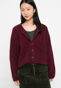 Mulher jovem com cabelo preto longo a usar um cardigã peludo de botão maroon sobre uma blusa verde escura e uma saia de veludo cotelê verde oliva com grandes bolsos.