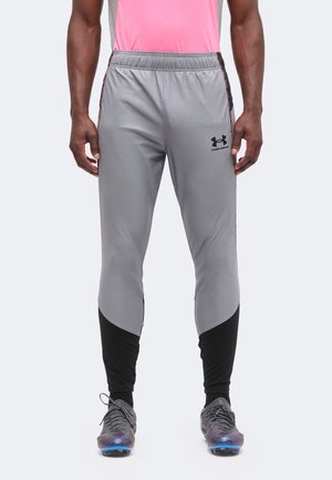 Homme portant un pantalon de sport Under Armour gris et noir et des crampons gris, debout devant un fond uni.