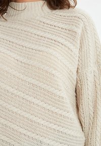 Pull en tricot crème clair avec un col montant, présentant des motifs côtelés diagonaux et une coupe ample, mettant en valeur une surface texturée.