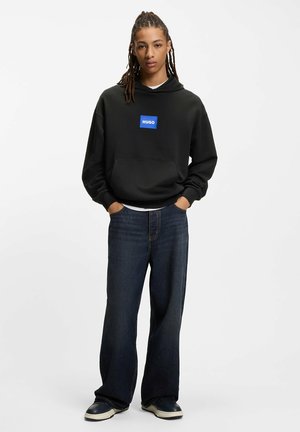 Black hoodie with a blue square logo reading "HUGO," paired with loose-fitting dark denim jeans and black and white shoes.