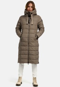 Knie-lange, gewatteerde pufferjas met capuchon in taupe, met een gestikte uitstraling, vooraan voorzien van een ritssluiting en trekkoordaccenten. Gecombineerd met witte broeken en beige laarzen.