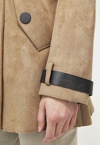 Brązowa zamszowa kurtka z czarnym skórzanym zapięciem na mankiet. Posiada zapięcie na guziki oraz dopasowany krój. Miękka struktura o matowym wykończeniu.