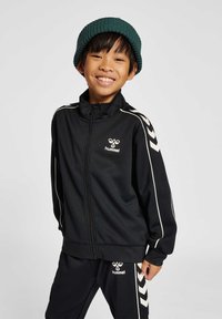 Chaqueta deportiva negra con cremallera frontal, detalles en blanco y logo. Combinada con pantalones negros que presentan un patrón de rayas diagonales. Gorro de punto verde.