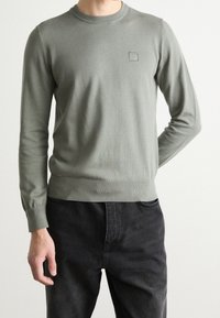 Mies, który ma na sobie jasnoszary sweter z długim rękawem i małym logo "BOSS" na piersi, oraz czarne dżinsy, stoi na tle jednolitego tła.