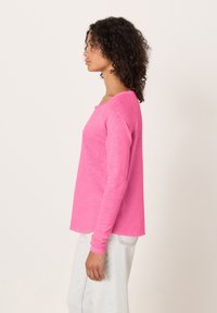 Ženska v profilu, oblečena v svetlo roza majico z dolgimi rokavi in svetle kavbojke, na enostavni svetli podlagi.