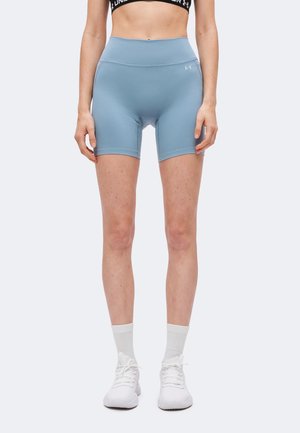 Femme portant un short cycliste taille haute bleu clair, des chaussettes blanches et des baskets blanches, debout devant un fond clair uni.