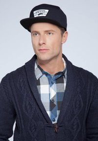 Cardigan lavorato a maglia blu navy con motivo a trecce, indossato sopra una camicia a quadri blu e grigia. Cappello nero con logo "VANS". Sfondo semplice.