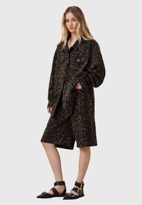 Veste en denim à imprimé léopard portée sur un short assorti, le tout dans des tons de marron et de noir. Sandales plates noires à lanières avec accents dorés.