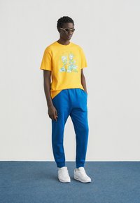 Κίτρινη βαμβακερή μπλούζα με γραφικό τύπωμα και πράσινες λεπτομέρειες, σε συνδυασμό με μπλε φόρμες και λευκά αθλητικά παπούτσια. Αξεσουάρ περιλαμβάνουν γυαλιά ηλίου.