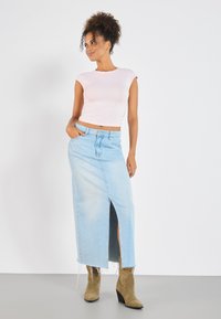 Un cropped top roz deschis, ajustat, cu mâneci scurte, asortat cu o fustă maxi din denim albastru deschis, cu talie înaltă, care are o fanta în față și o margine deteriorată.