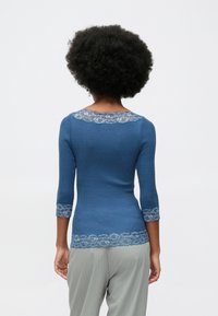 Göndör hajú nő, aki kék bordás felsőt visel, csipke díszítéssel a ujjaknál, a nyakán és az alján, világosszürke csíkos nadrággal, hátrafordulva.