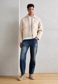 Veste beige texturée avec deux poches poitrine, associée à un jean bleu droit et des baskets beiges, sur un fond neutre.