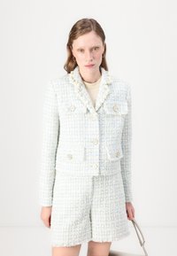 Šviesiai mėlynos ir baltos spalvų tweed dviejų dalių komplektas, kuriame yra trumpa striukė su auksiniais mygtukais ir pakraščiu su kutais, derinama su šortais.