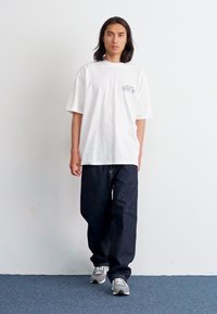 White oversized t-shirt with a small graphic print on the left chest, paired with dark blue loose-fitting jeans and light gray sneakers.