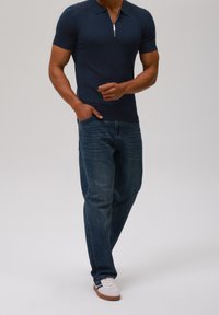 Marinblå kortärmad polo med ribbad textur och dragkedja i kragen, i kombination med blå denimjeans och vita sneakers med bruna sulor.