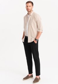 Camisa de botonadura ligera color beige sobre una camiseta blanca, combinada con pantalones negros y zapatos gris claro de tipo slip-on. Presenta un bolsillo en el pecho y un reloj.