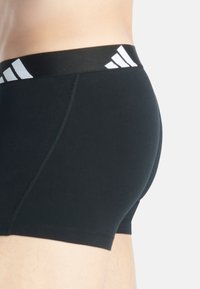 Svarta boxertrosor med slät textur, med en bred svart midjeband som har vit tre-stripes branding, visad från sidan.