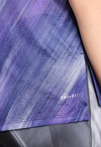 Close-up van een paars patroon Dri-FIT sportshirt gedragen over grijze legging, met nadruk op de stoftextuur en stikdetails.