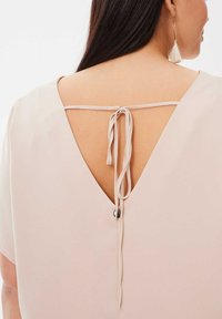 Lichtbeige top met een diepe V-rug en een gestrikte sluiting. De zachte textuur en korte mouwen versterken het minimalistische ontwerp.